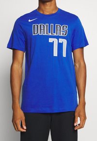 Uomo che indossa una maglietta blu Nike del Dallas con il numero 77, abbinata a pantaloni corti neri, in piedi davanti a uno sfondo bianco semplice.