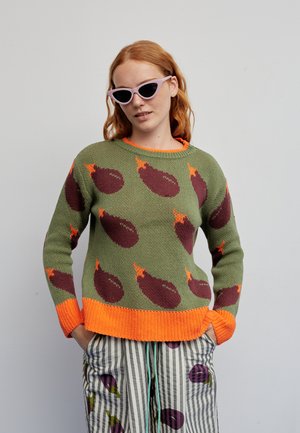 Pull en maille vert olive avec motifs aubergine violet et accents orange. Le modèle porte des lunettes de soleil œil de chat violet clair et un pantalon rayé.