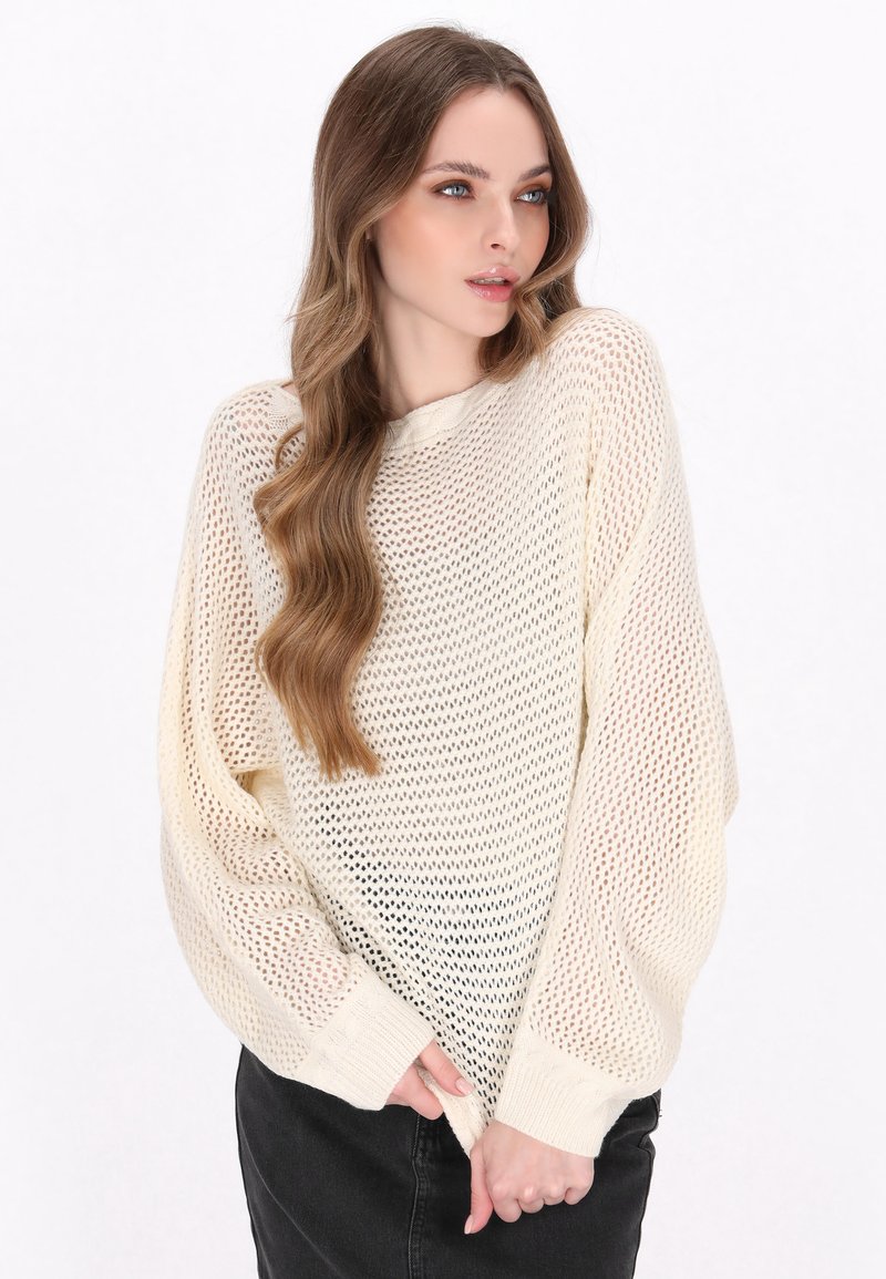 Lichte, oversized crème gebreide trui met een losse meshstructuur. Voorzien van lange mouwen en geribbelde manchetten, gecombineerd met zwarte broek.