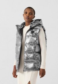 Smanicato imbottito argento con una texture lucida e trapuntata e un cappuccio. Presenta una chiusura con zip e tasche laterali, indossato sopra un capo di colore chiaro.