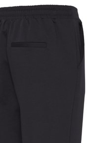 Gros plan sur un pantalon noir montrant une taille élastique, une poche arrière passepoilée et une partie de la couture d’une poche latérale.