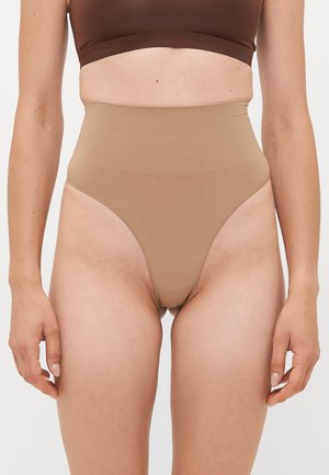 Woman wearing high-waisted beige shaping underwear and a brown cropped top, standing with arms relaxed at sides.
