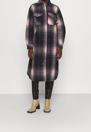 Lange geruite jas in roze, zwart en paars met knopen aan de voorkant en borstzakken, gedragen met zwarte broek en beige enkellaarzen.