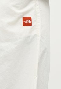 Λευκό ύφασμα με ένα μικρό κόκκινο τετράγωνο μπαλώμα που φέρει το λευκό λογότυπο "The North Face" ραμμένο κοντά σε μια ραφή.