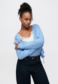 Ανοιχτό μπλε πλεκτή ζακέτα με ζώνη στη μέση και μακριά μανίκια, φορεμένη πάνω από λευκό τοπ με δαντέλα και σκούρο τζιν.