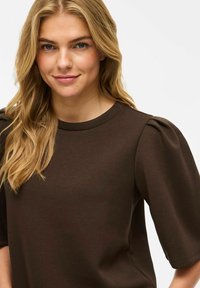 Brūns īsās piedurknes topiņš ar noapaļotu kakla izgriezumu un puffed plecu dizainu, izgatavots no gludas, vieglas auduma.