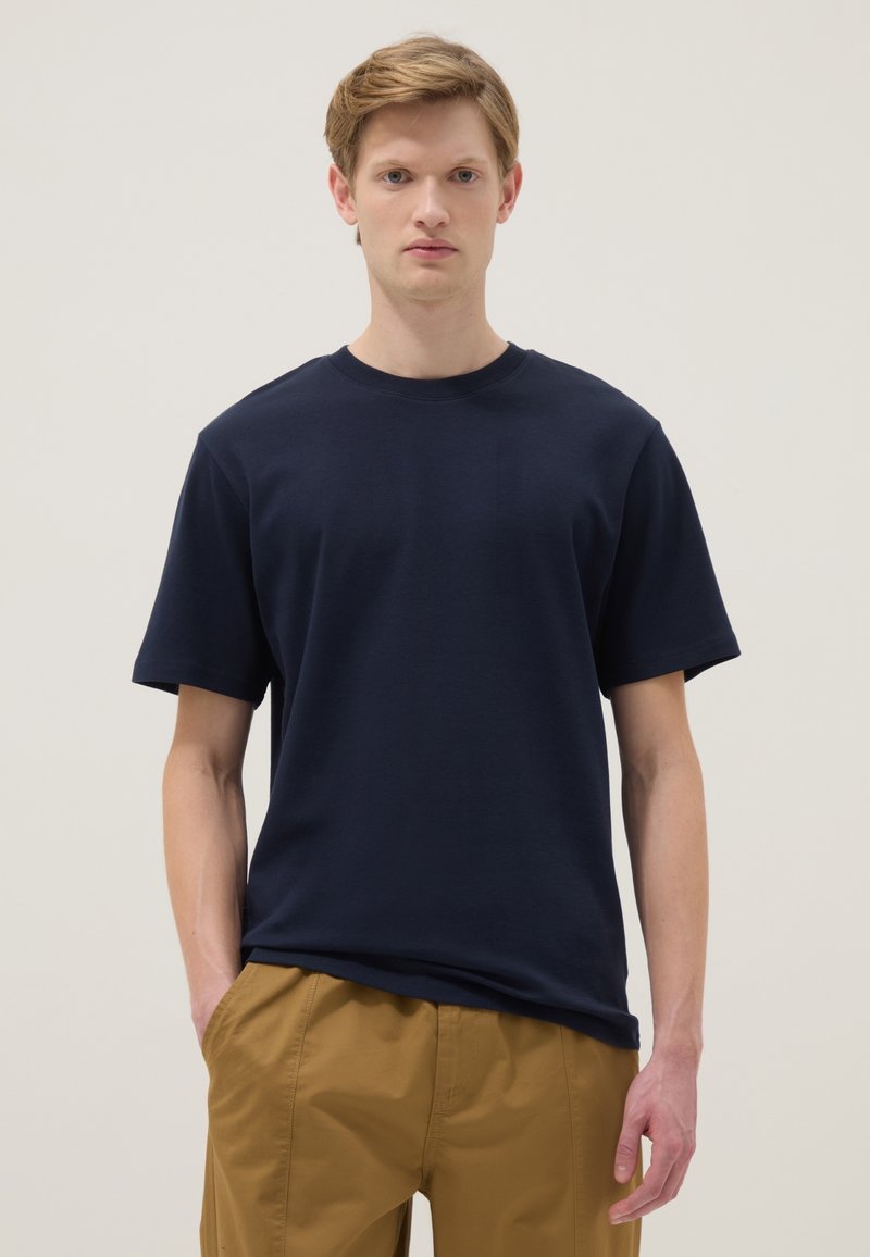 Joven con cabello castaño claro, vistiendo una camiseta de manga corta azul marino y pantalones color mostaza, de pie con una mano en el bolsillo.