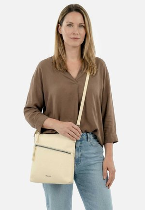 Vrouw draagt een bruin shirt en spijkerbroek, met een crème schoudertas met een voorvak met rits tegen een effen achtergrond.