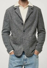 Blazer in lana grigia con colletto a revers, dotato di due tasche frontali e due bottoni. Indossato sopra un maglione chiaro e lavorato.