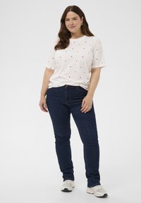 Weißes T-Shirt mit verstreuten kleinen roten Herzmotiven, kombiniert mit dunkelblauen Jeans. Das Outfit zeichnet sich durch eine lockere Passform und kurze Ärmel aus.