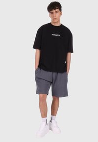 Schwarzes oversized T-Shirt mit weißem Logo, kombiniert mit grauen strukturierten Shorts. Modell trägt weiße Sneakers mit Branding und weiße Socken.