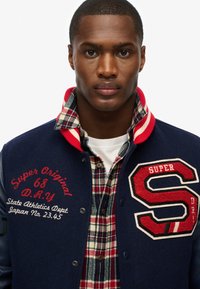 Chaqueta de varsity azul con acentos en rojo y blanco, que presenta un gran parche con "S", texto bordado y una camisa de cuadros debajo.