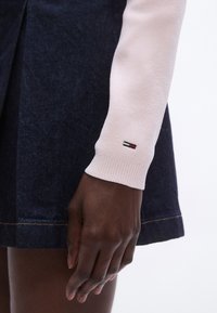 Top rosa chiaro a maniche lunghe con polsini a coste; indossato sopra una gonna in denim scuro con cuciture visibili e una leggera svasatura. Dettaglio logo sulla manica.