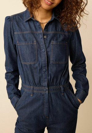 Femme portant une combinaison en denim bleu foncé à manches longues, fermeture par boutons sur le devant, poches poitrine et mains dans les poches latérales.