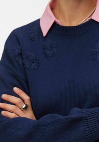 Personne portant un pull en maille bleu marine avec des appliqués floraux sur une chemise rose à col, les bras croisés, et une bague en argent à la main gauche.