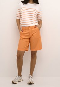 Oranžové bavlněné kraťasy s kapsami, v kombinaci s bílým topem s oranžovými vodorovnými pruhy. Model má na sobě světlé tenisky.