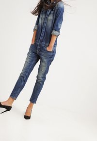 Denimjumpers med en ljus till mörk tvättgradient, långärmade uppvikt, två bröstfickor, slitna detaljer och avsmalnande ben. Svarta högklackade skor.