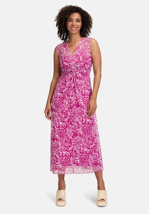 Ein langes, pinkes Blumenkleid mit V-Ausschnitt, Bindegürtel und kunstvollen weißen Mustern. Das Model trägt beige Plattformschuhe.