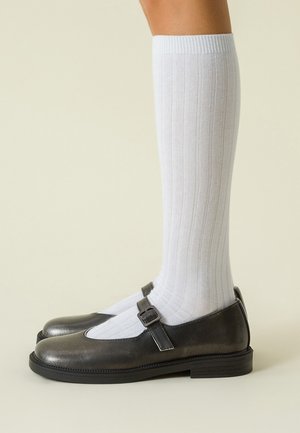 Jambes portant des chaussettes blanches à côtes montant jusqu'aux genoux et des chaussures Mary Jane en cuir noir avec des sangles à boucle, se tenant sur un fond neutre.