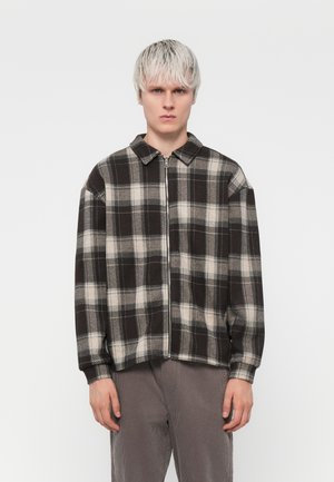 Zwarte en beige geruite zip-up jas met een kraag, losse pasvorm en zachte textuur; gedragen over grijze ribfluwelen broek.