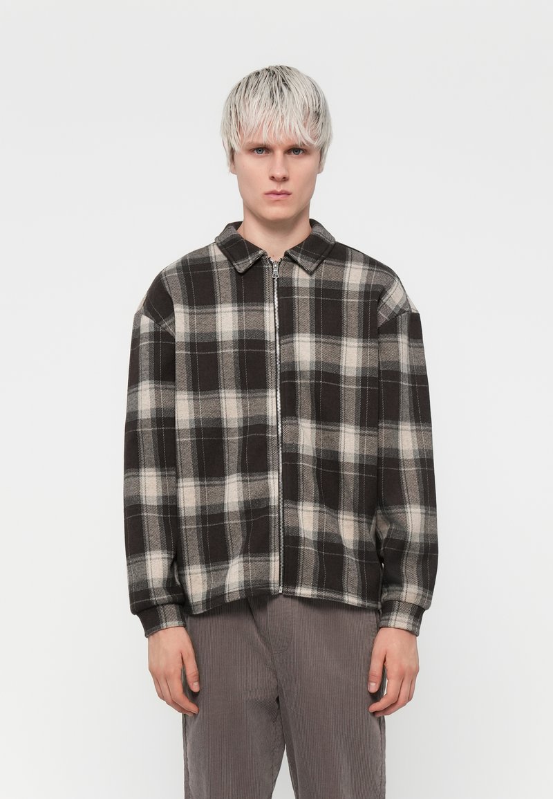 Black and beige plaid zip-up jacket with a collared neck, loose fit, and soft texture; worn over grey corduroy trousers.