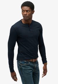 Dunkelblaues Langarm-Henley-Shirt mit Knopfleiste, dezentem eingestanztem Logo und gerippten Bündchen. Getragen mit hellblauen Jeans und brauem Gürtel.