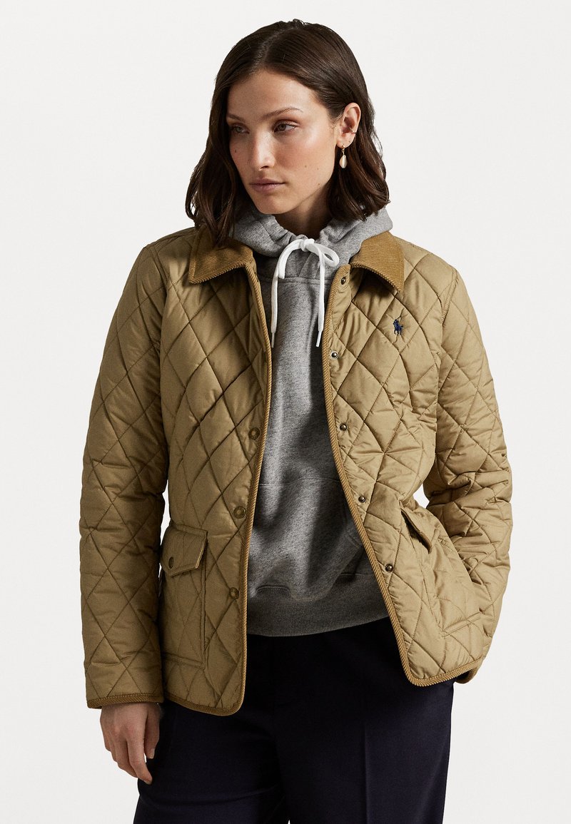 Gequiltede bruine jas met een ribbelkraag, voorzien van een voorste drukknoopsluiting en zijzakken. Gedragen over een grijze hoodie.