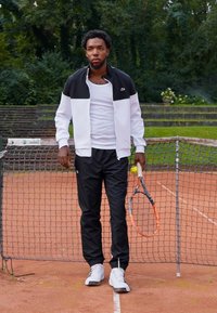 Jachetă de tenis albă și neagră, pantaloni negri și adidași albi. Ținând o rachetă de tenis și o minge pe un fundal de teren de zgură.
