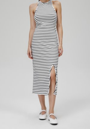Vestido midi de rayas en blanco y negro, con un cuello alto, diseño sin mangas y una abertura lateral. Combinado con zapatos blancos tipo slip-on.