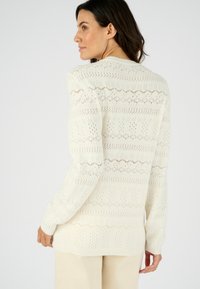 Pull en maille blanche avec des motifs ajourés et des designs géométriques, à manches longues et avec des bords côtelés. Texture subtile et apparence légère.