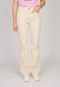 Jeans beiges évasés avec taille haute, poches avant et une légère brillance ; associés à des baskets blanches, créant un look décontracté.
