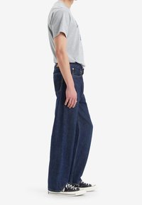 Homme debout de profil, portant un t-shirt gris, un jean large bleu foncé et des baskets basses noires avec des lacets blancs.
