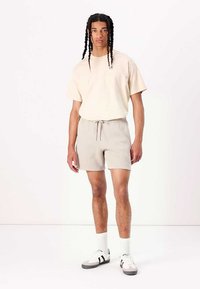 Camiseta de algodón beige combinada con pantalones cortos grises claros de cordón. Zapatillas casuales con detalles en negro y blanco, de pie sobre un fondo sencillo.