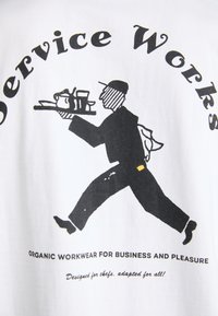 White T-shirt featuring a black graphic of a person carrying a tray with cups. Text reads "Service Works" and "Organic workwear for business and pleasure."