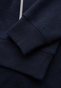 Détail de la manche et du poignet d’un sweat-shirt bleu marine avec une texture côtelée visible et une fermeture éclair argentée partiellement visible sur le côté.