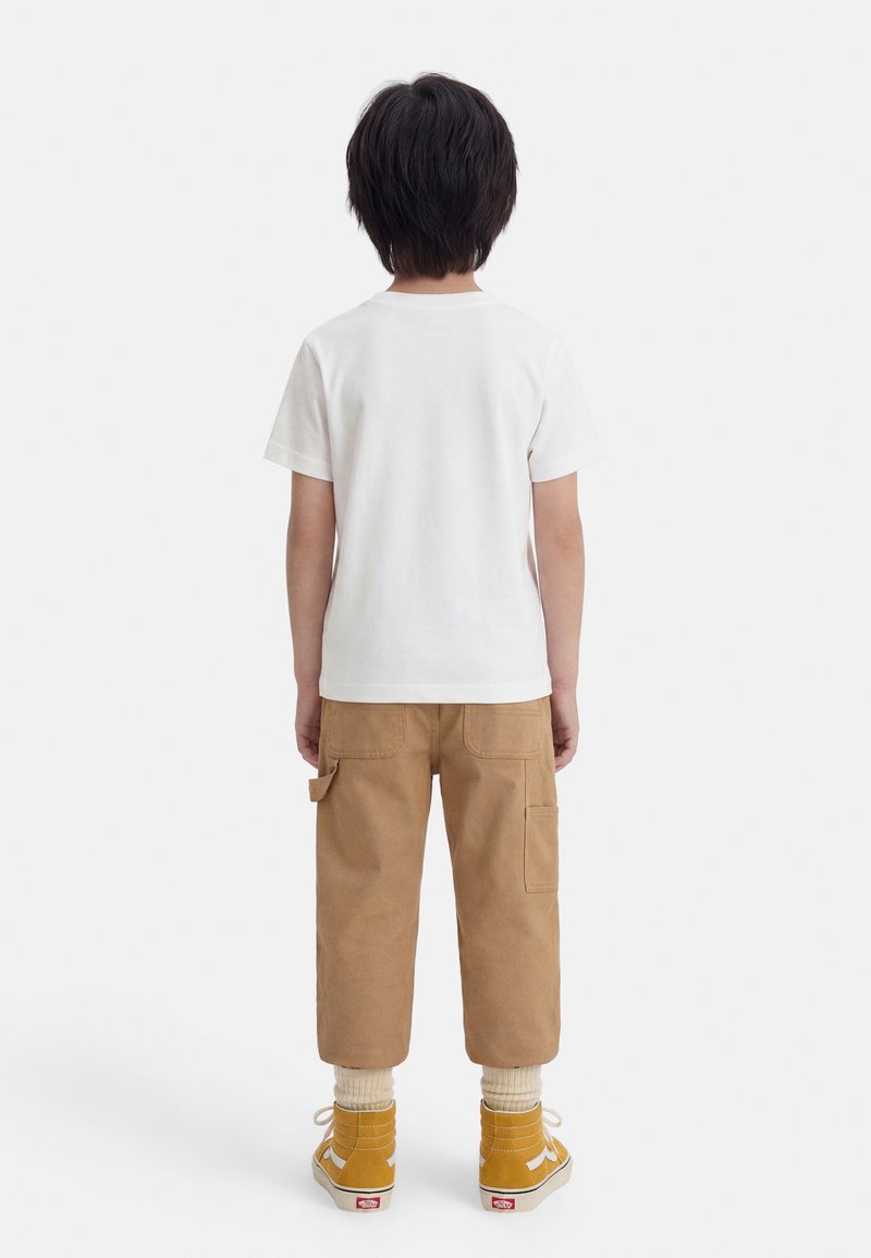 Kind staat met de rug naar de kijker, draagt een gewoon wit t-shirt, bruine cropped broek met zakken, crèmewitte sokken en gele hoge sneakers.