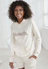 Cremefarbenes Hoodie mit einer Fronttasche und Textlogo. Kurze cremefarbene Shorts mit passendem Logo. Glatte Textur und lockere Passform.