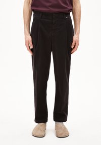 Zwarte, geplooide katoenen broek met een rechte pijpvorm, voorzien van riemlussen en zijzakken, gecombineerd met beige instapschoenen.