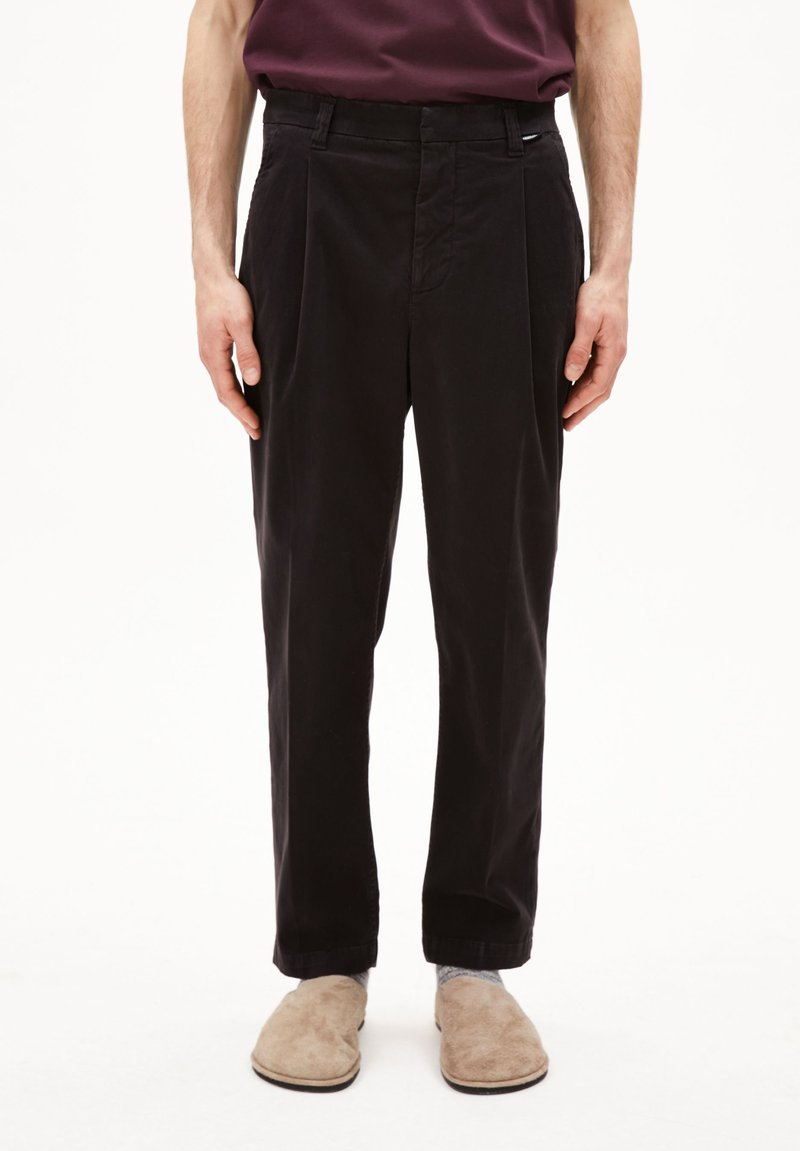 Zwarte, geplooide katoenen broek met een rechte pijpvorm, voorzien van riemlussen en zijzakken, gecombineerd met beige instapschoenen.