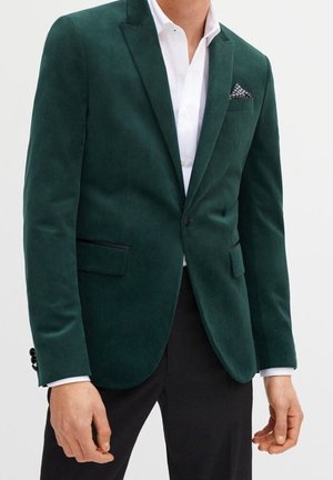 Mies, joka pukeutunut tummanvihreään samettiseen bleiseriin, jossa on lovikäänteet, valkoinen juhlapaita, mustat housut ja pilkullinen taskuliina.