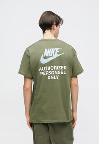 Ελαιόχρωμο υπερμεγέθες T-shirt από βαμβάκι. Διαθέτει λευκό λογότυπο "NIKE" και κείμενο "ΜΟΝΟ ΕΞΟΥΣΙΟΔΟΤΗΜΕΝΟ ΠΡΟΣΩΠΙΚΟ" στην πλάτη. Κοντά μα sleeves.