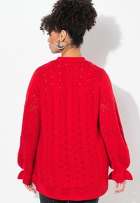 Roter gestrickter Pullover mit einem strukturierten Muster, lockerer Passform, Ballonärmel und Durchbrüche entlang des Rückens und an den Seiten.