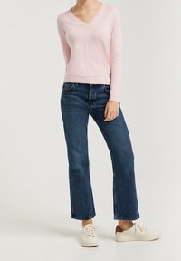 Woman wearing light pink V-neck sweater, dark blue straight-leg jeans, and white sneakers standing on light background.