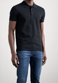 Man i svart kortärmad polo t-shirt och blå jeans som står mot en enkel, ljus bakgrund.