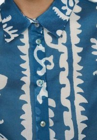 Chemise à motifs bleus et blancs, avec col, devant boutonné avec des boutons ronds, et des dessins distincts en forme de vagues et abstraits.