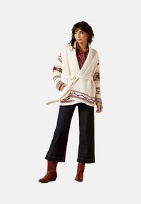 Weißer gestrickter Wickelcardigan mit burgunderfarbenen Mustern, weiten Ärmeln und einem gebundenen Gürtel, kombiniert mit dunklen Schlagjeans und roten Ankle Boots.
