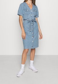 Vaaleansininen farkkukankainen paidatidress, jossa on lyhyet puffihihat, vyötäröllä vyö, kaksi rintataskua ja polvenpituinen malli. Valkoiset tennarit jalassa.