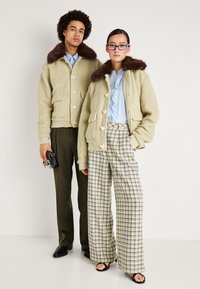 Bársonyos bézs kötött kabátok barna szőrmés gallérral, gombos elülső résszel és oldalsó zsebekkel; csíkos nadrággal és mintás, bő szárú nadrággal párosítva.