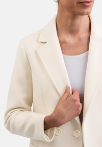 Blazer bianco con una vestibilità strutturata, dotato di rever a punta, due bottoni e tessuto liscio. Indossato sopra una maglietta chiara, che mette in risalto un design pulito.