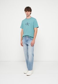 Světle tyrkysové bavlněné tričko s tmavým logem, doplněné světlými džínami. Model stojí ve bílých teniskách před neutrálním pozadím.